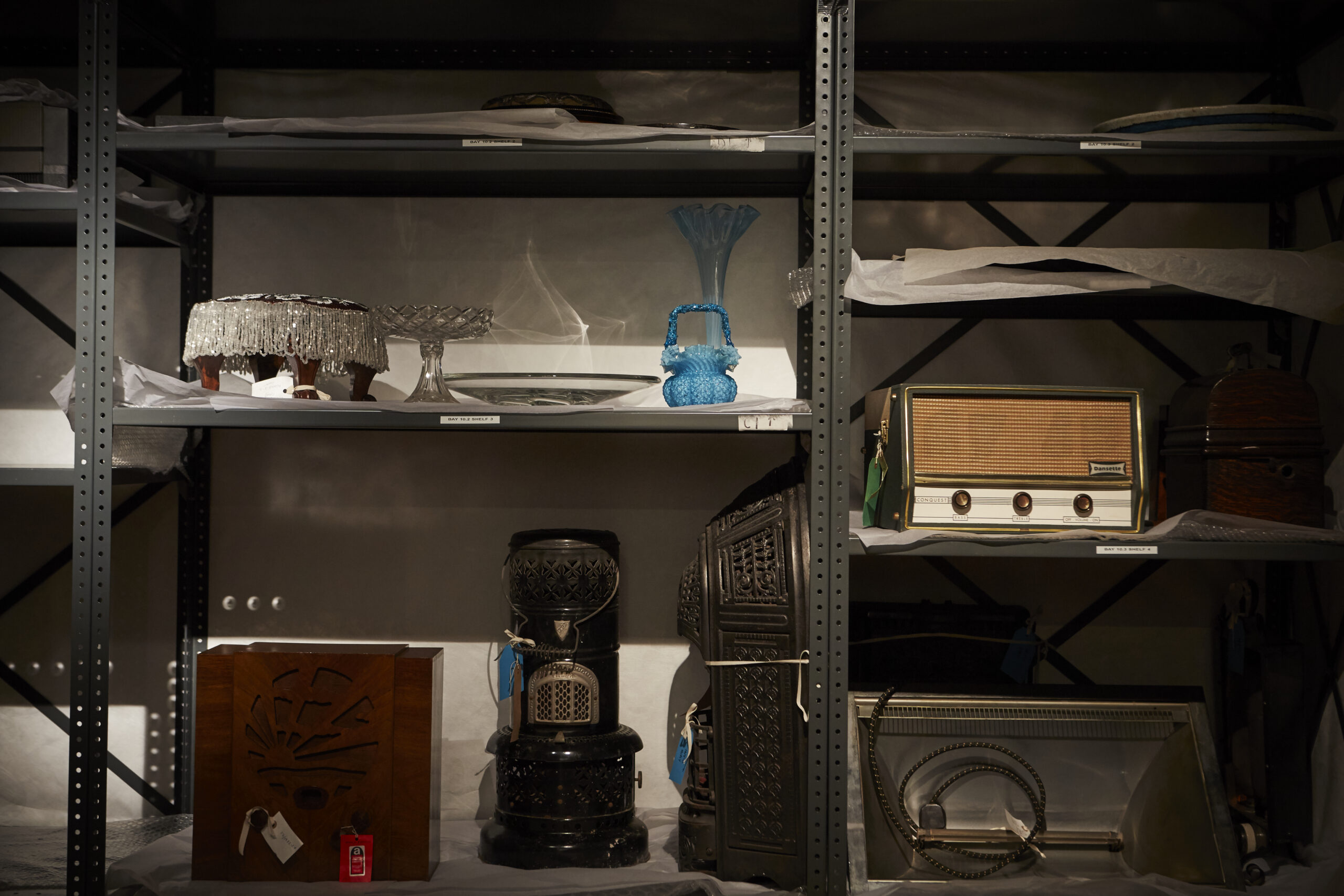 A dimly lit steel shelving unit lined with white tissue paper and labelled with small white stickers. The shelves hold stored objects including a shallow glass platter, plates, a blue glass basket, a tall flared blue glass vase, a pale yellow vintage radio, a wooden radio cabinet, and more.
