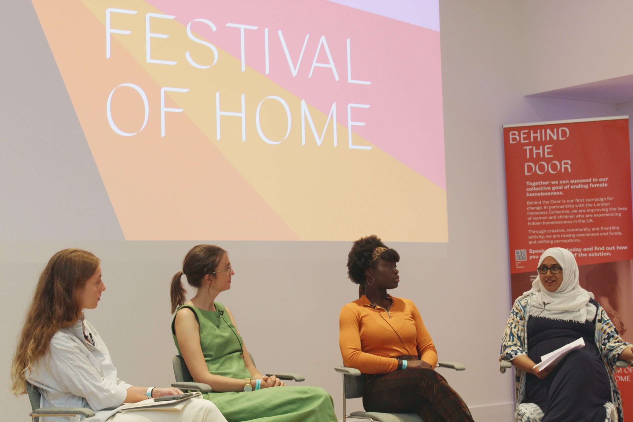 Four women on a panel discussion with colorful backdrop reading 'Festival of Home' and "Behind the Door" campaign banner about female homelessness.