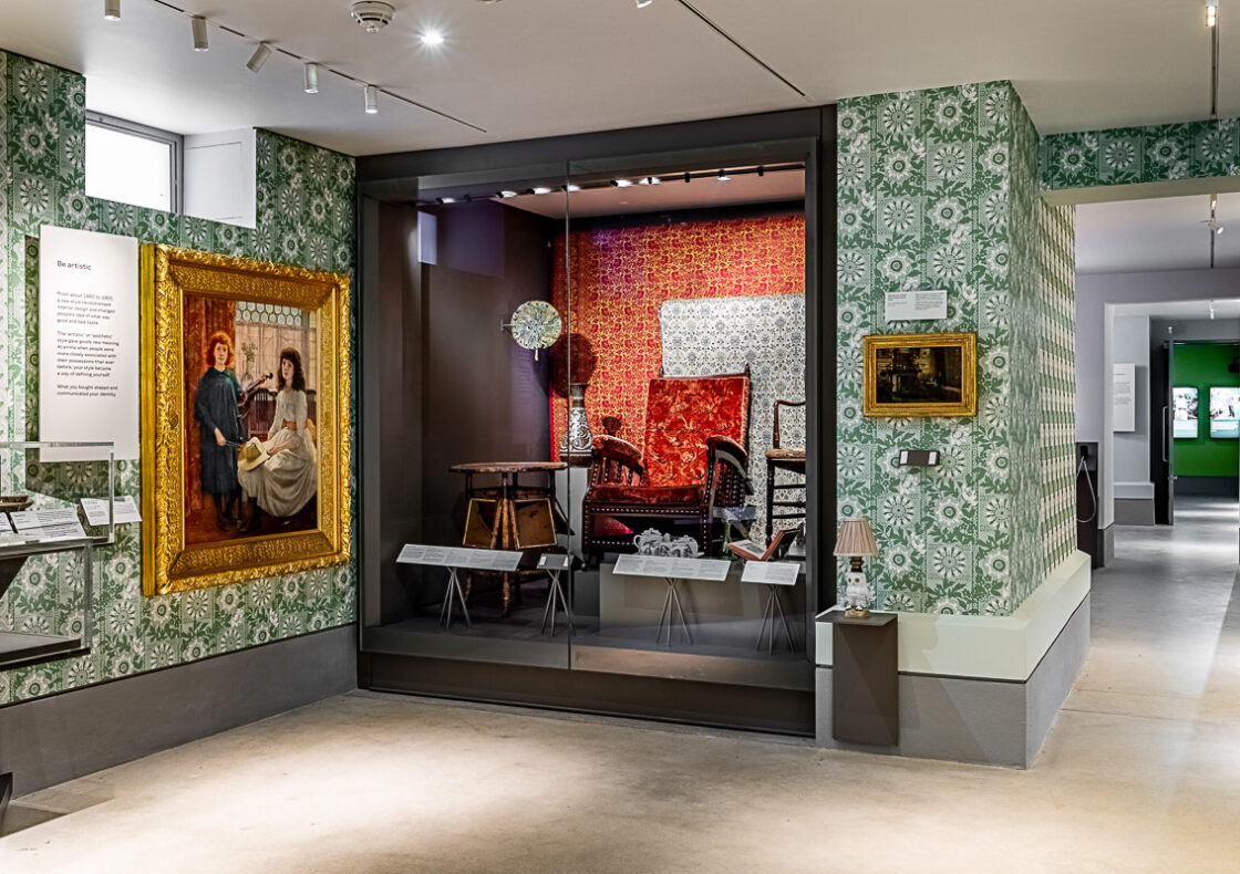 Two framed paintings hang on patterned green and wallpaper; in the middle is a glass case displaying period chairs with ornate wallpaper.