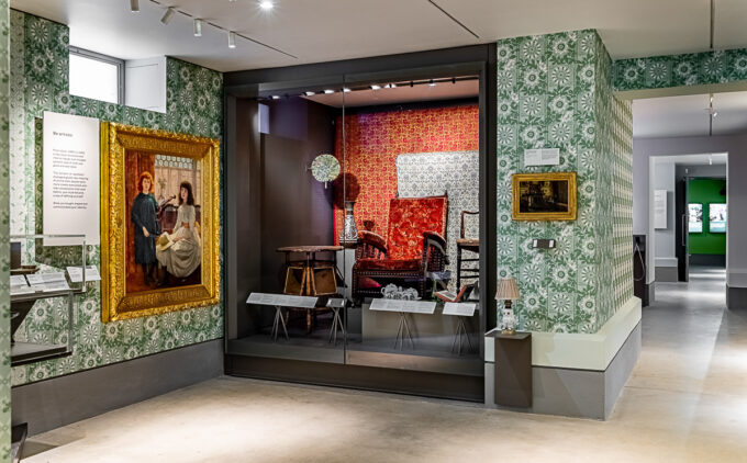 Two framed paintings hang on patterned green and wallpaper; in the middle is a glass case displaying period chairs with ornate wallpaper.