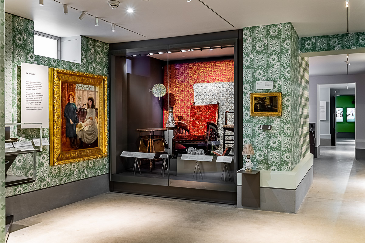 Two framed paintings hang on patterned green and wallpaper; in the middle is a glass case displaying period chairs with ornate wallpaper.