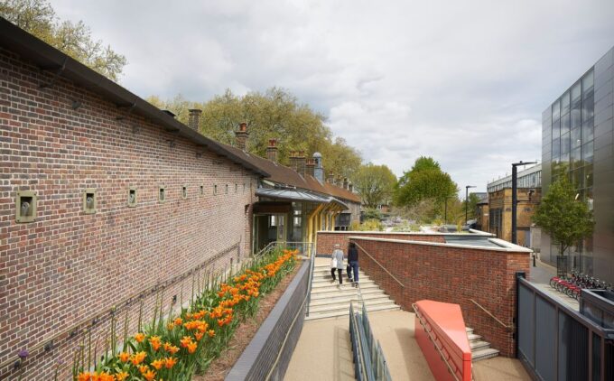 A step-free ramp with handrails leads to the Hoxton entrance of the Museum, bordered by a bed of orange flowers, while two visitors climb adjacent stairs.