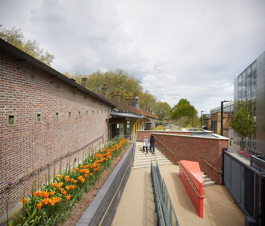 A step-free ramp with handrails leads to the Hoxton entrance of the Museum, bordered by a bed of orange flowers, while two visitors climb adjacent stairs.