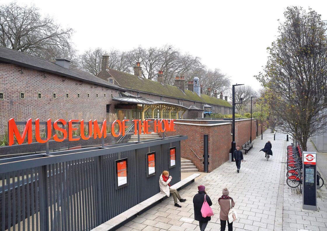 A street-level view of the Museum of the Home entrance: a long dark metal fence topped by bright orange capital letters spelling “Museum of the Home” lines a paved pedestrian alley. Behind it, a row of moss-topped brick almshouses with chimneys stretches beneath bare winter trees, while a handful of pedestrians stroll along the path.