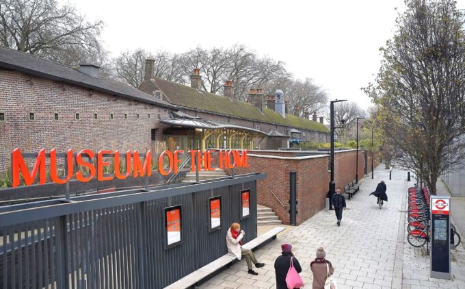 A street-level view of the Museum of the Home entrance: a long dark metal fence topped by bright orange capital letters spelling “Museum of the Home” lines a paved pedestrian alley. Behind it, a row of moss-topped brick almshouses with chimneys stretches beneath bare winter trees, while a handful of pedestrians stroll along the path.