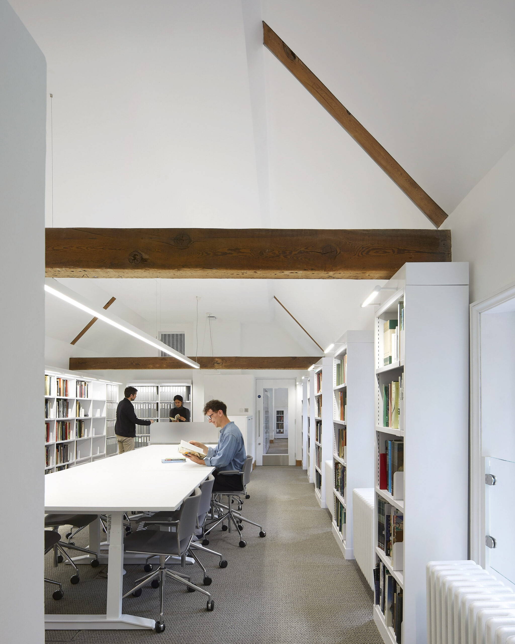 A person reads at a long white table in a Collection Library, a bright reading room with full bookshelves and exposed wooden beams on the roof, while two people chat by the shelves and two more converse in the background.