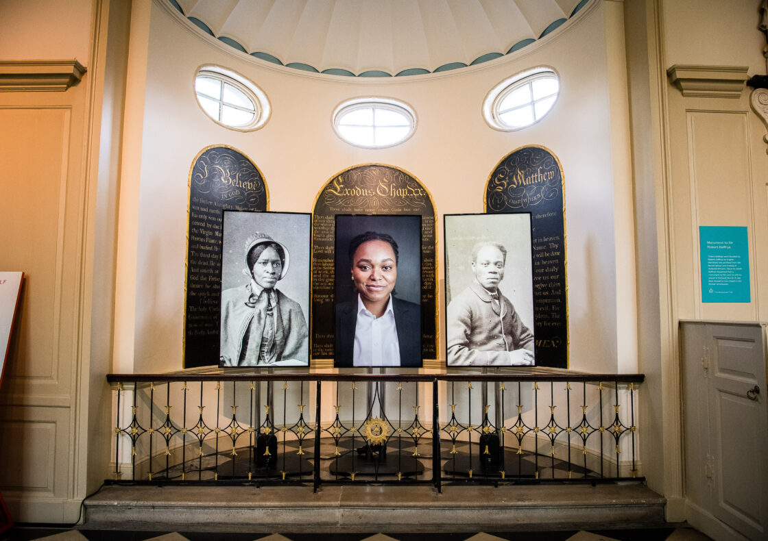 Portrait gallery display with three framed images, two historical Black and white photographs flanking a contemporary colour portrait, in front of decorative panels with oval windows above.