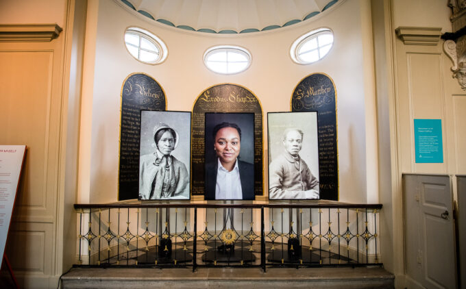 Portrait gallery display with three framed images, two historical Black and white photographs flanking a contemporary colour portrait, in front of decorative panels with oval windows above.