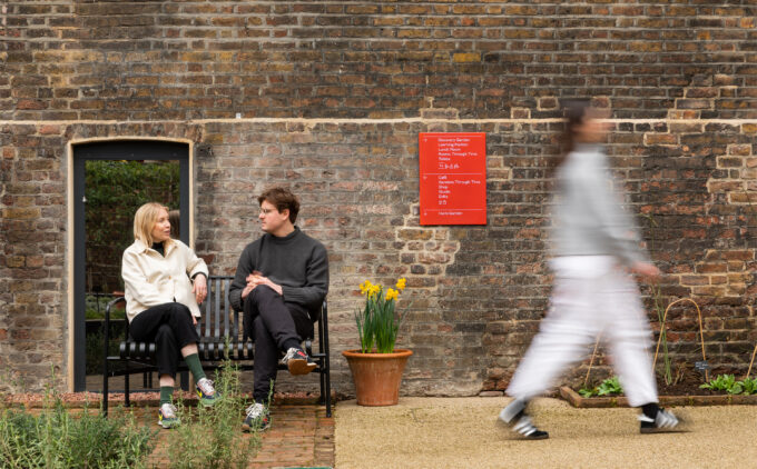 Two people sit on a bench outside, engaged in conversation against a brick wall. A blurred passerby walks by. A potted plant and a red sign are visible.