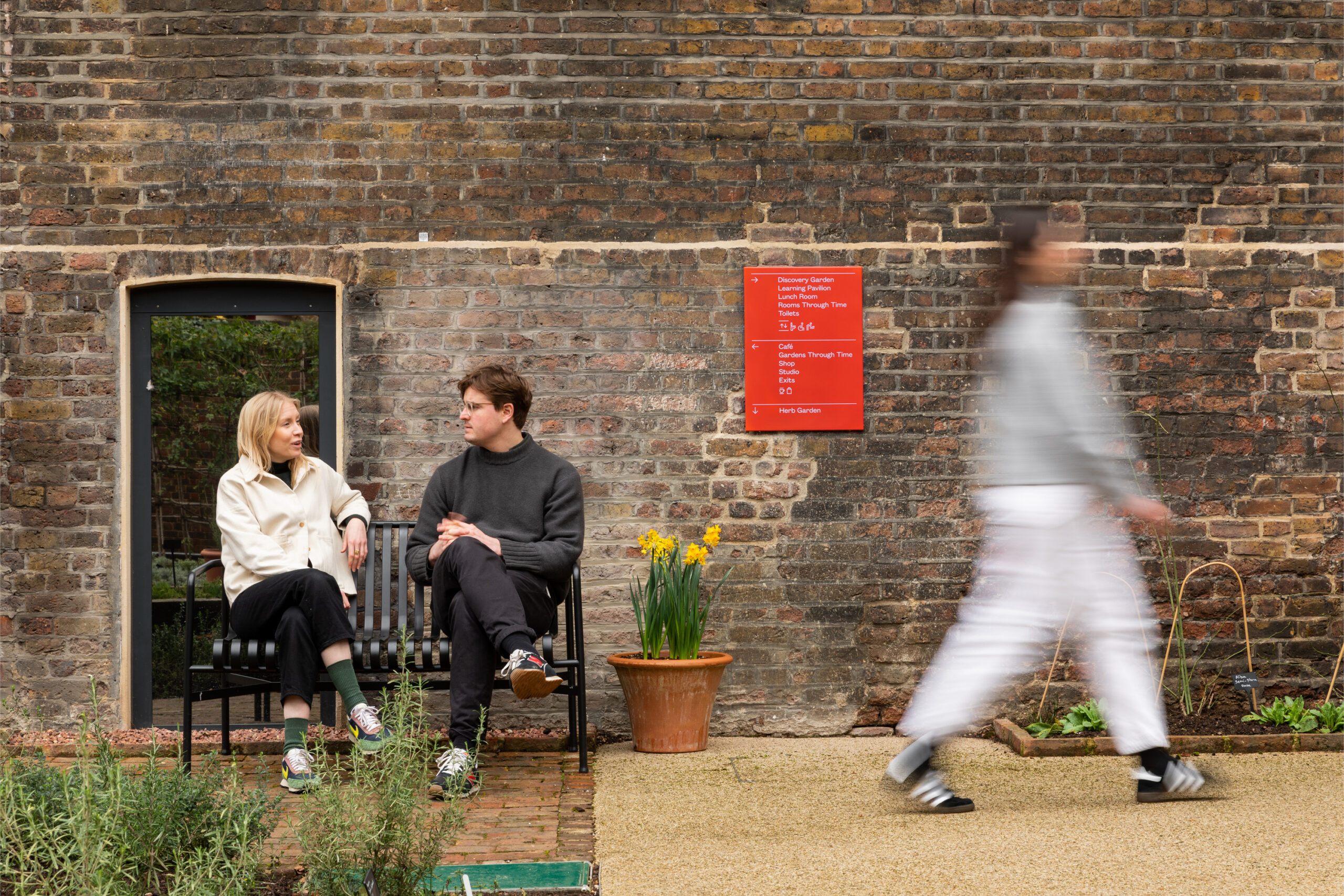 Two people sit on a bench outside, engaged in conversation against a brick wall. A blurred passerby walks by. A potted plant and a red sign are visible.