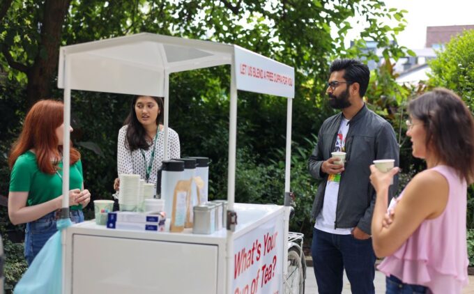 A woman serves drinks from a white cart labeled 'What's Your Cup of Tea?' while people gather around in an outdoor setting
