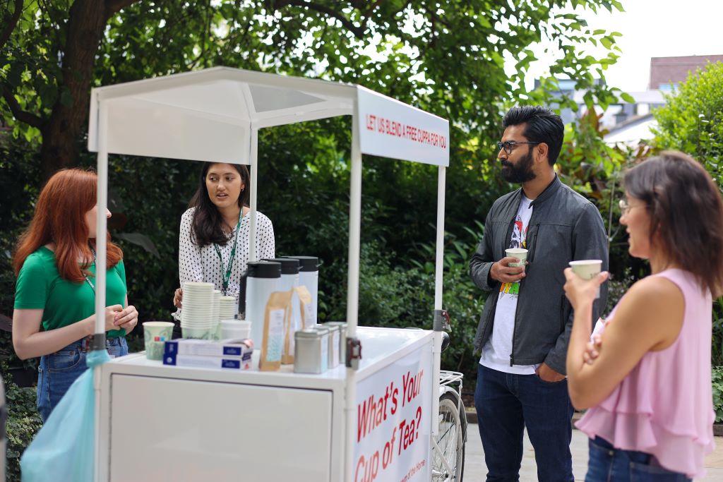 A woman serves drinks from a white cart labeled 'What's Your Cup of Tea?' while people gather around in an outdoor setting
