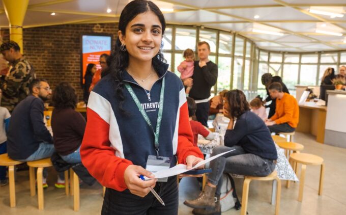 A volunteer wearing a green “Volunteer” lanyard smiles while holding a clipboard and pen in a busy gallery space where families sit at tables behind her.