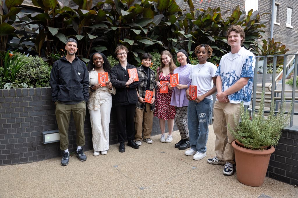 A group of young people pose outdoors holding orange books, smiling at the camera near leafy plants.