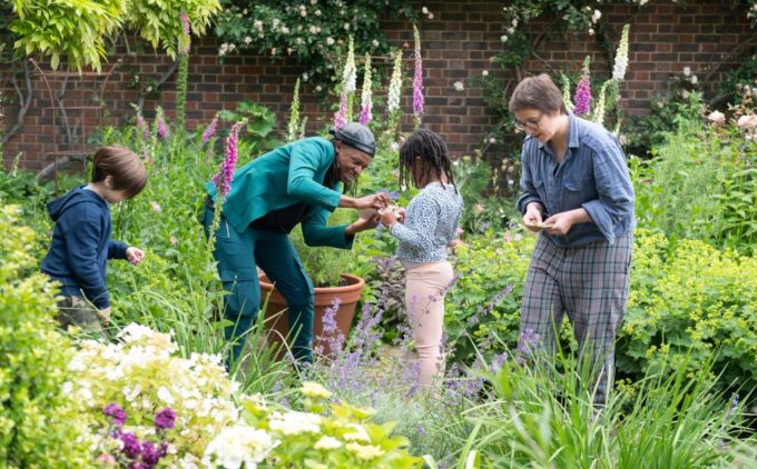 Two adults and two children pick and examine flowers in a lush garden with magenta and white foxgloves and a brick wall in the background.