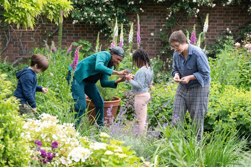 Two adults and two children pick and examine flowers in a lush garden with magenta and white foxgloves and a brick wall in the background.