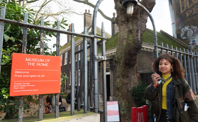 By the Kingsland Road entrance fence, a staff member holds a two-way radio while standing to the right of the Museum's red orange "Welcome” sign and iron railings.