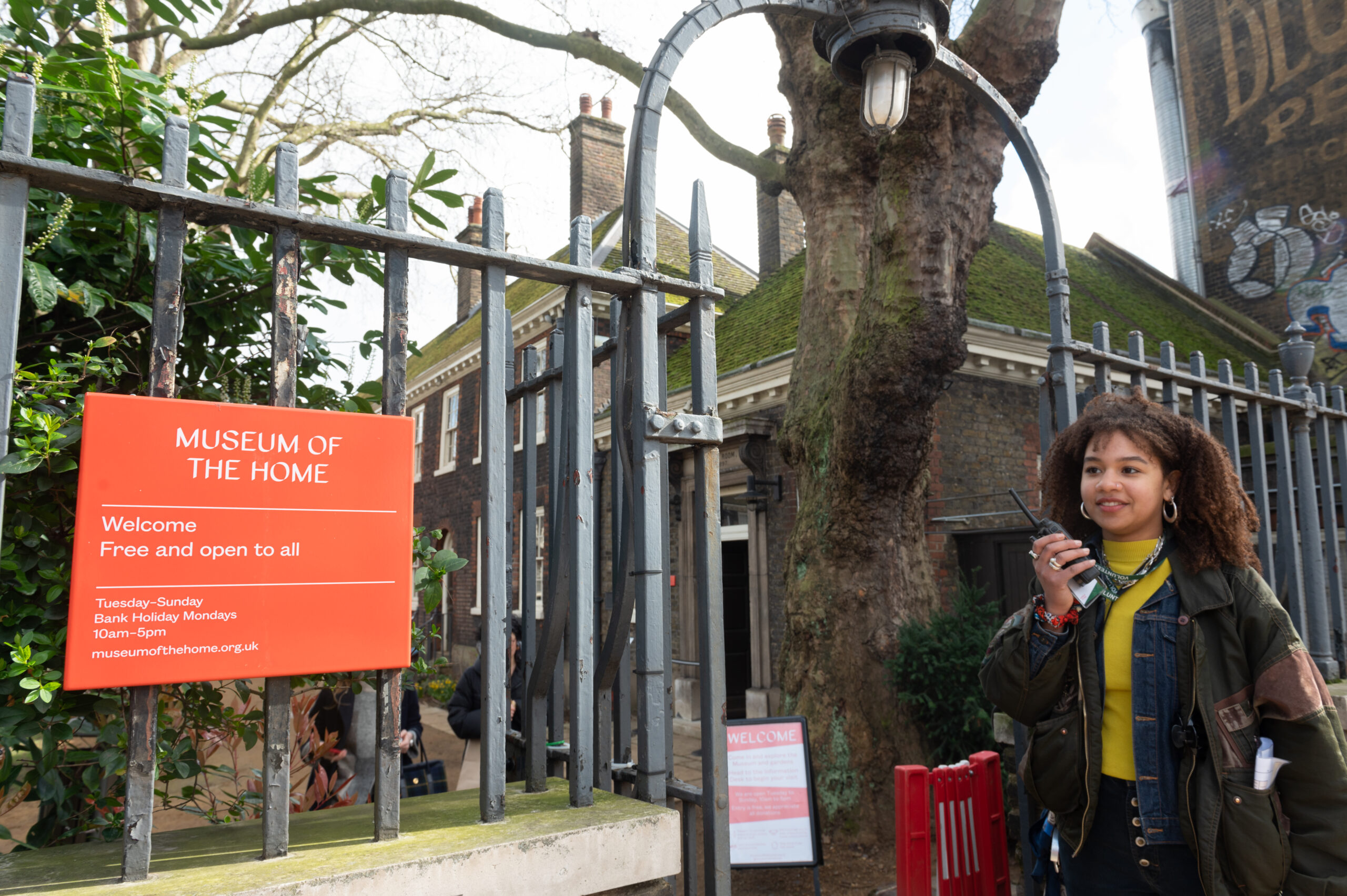 By the Kingsland Road entrance fence, a staff member holds a two-way radio while standing to the right of the Museum's red orange "Welcome” sign and iron railings.