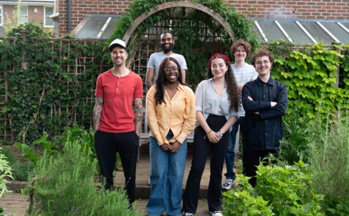 A group of six young people stands in front of a wooden garden arch surrounded by lush greenery, smiling as they face the camera.