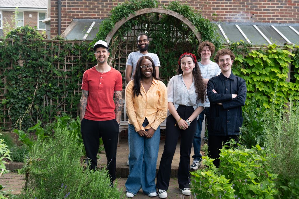 A group of six young people stands in front of a wooden garden arch surrounded by lush greenery, smiling as they face the camera.