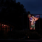 A colourful neon light display of a winged figure with a trident, a partially illuminated building is to the left, with dark trees silhouetted against the night sky in the background.