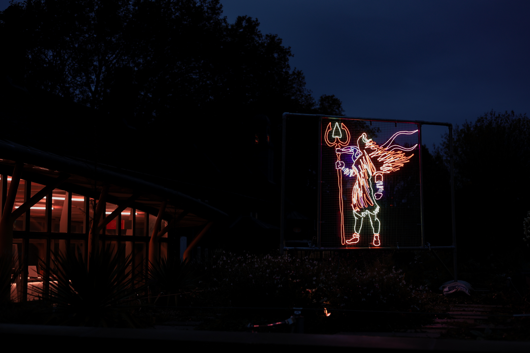 A colourful neon light display of a winged figure with a trident, a partially illuminated building is to the left, with dark trees silhouetted against the night sky in the background.