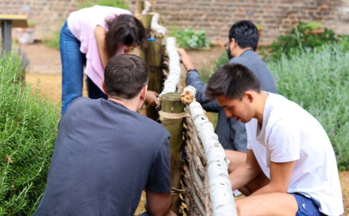 Two volunteers on each side of a rustic garden fence weave slender branches into upright posts amid surrounding greenery.