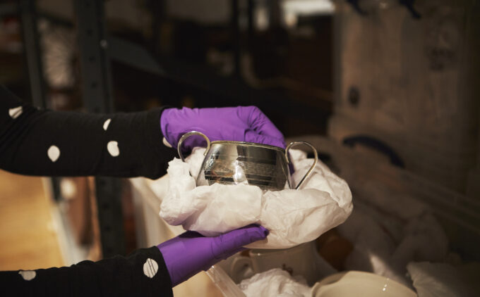 In the Museum’s collection store, gloved hands lift a silver vessel with handles wrapped in tissue paper.
