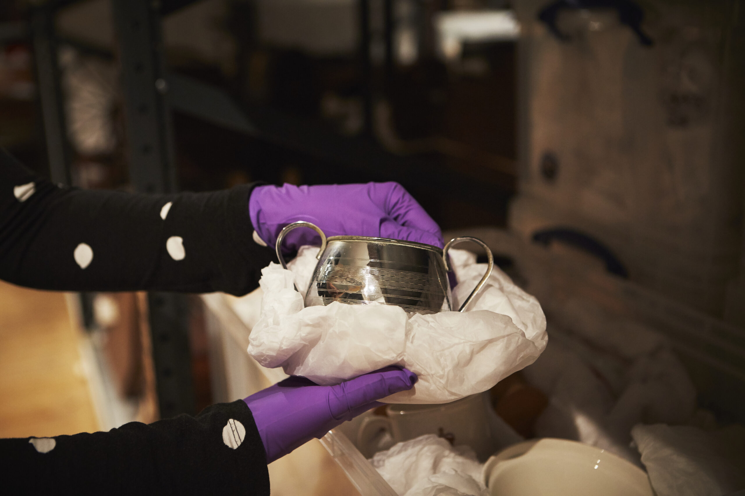 In the Museum’s collection store, gloved hands lift a silver vessel with handles wrapped in tissue paper.