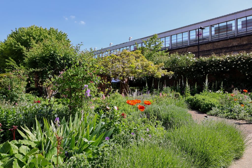 A garden filled with greenery and flowers and a small brick path. In the background is an overground train station.