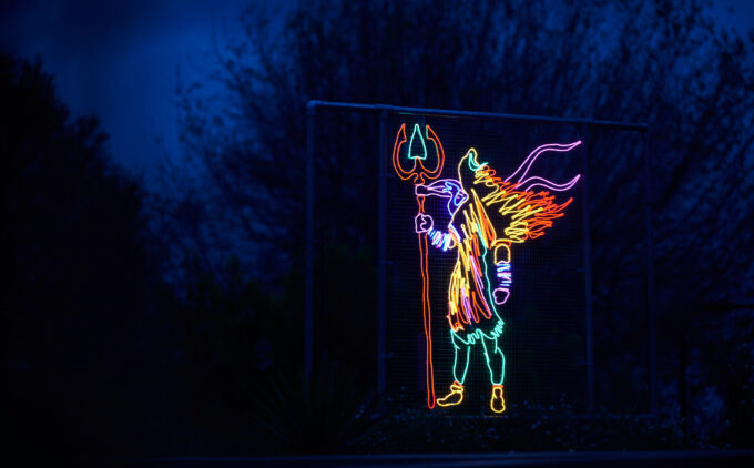 A neon outline of various colours of Shiva the Destroyer installation against a dark blue sky with some tree branches in the background, holding a red-and-green trident; the body is rendered in pink, yellow, and blue.