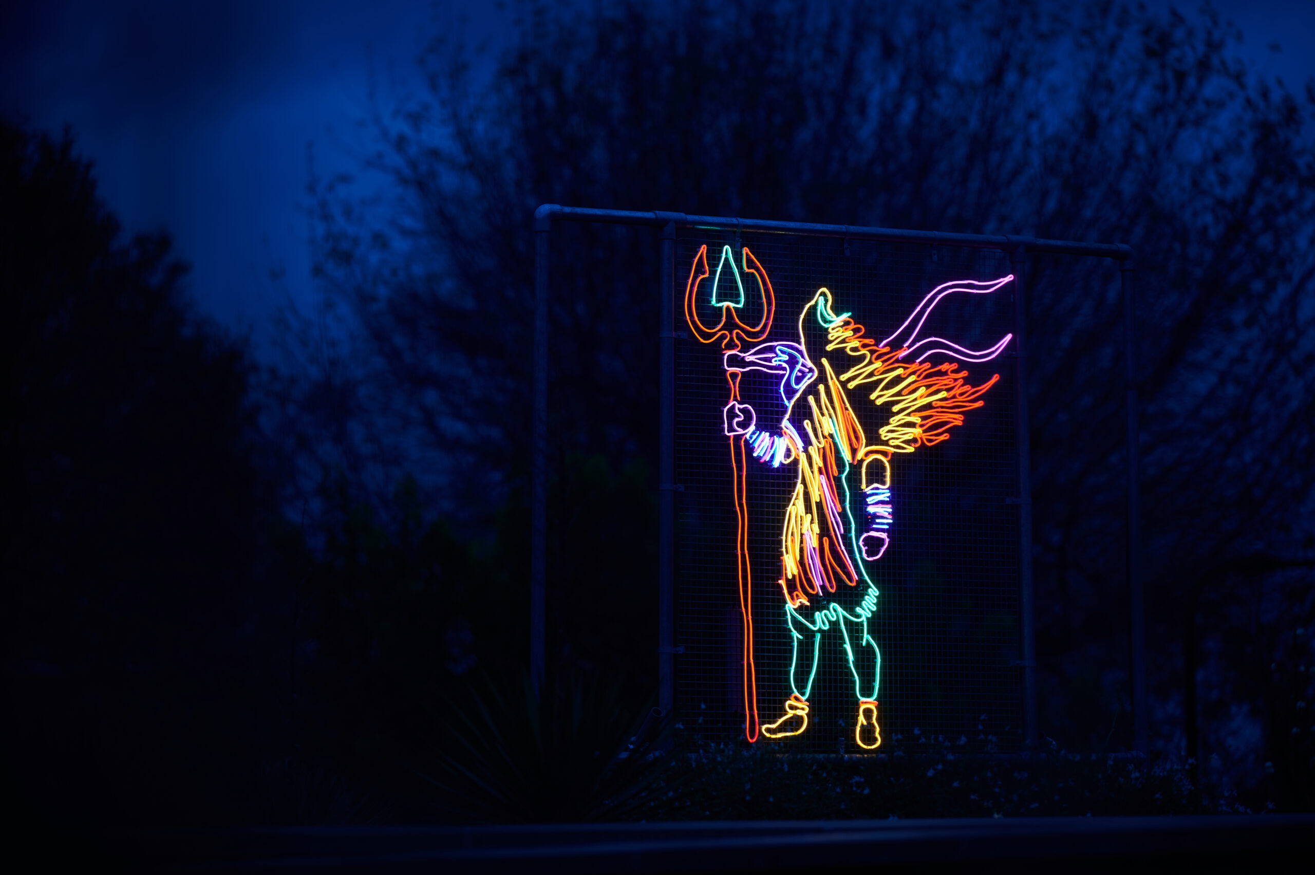 A neon outline of various colours of Shiva the Destroyer installation against a dark blue sky with some tree branches in the background, holding a red-and-green trident; the body is rendered in pink, yellow, and blue.