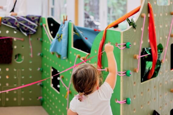 A young child pulls a pink ribbon towards a bright green pegboard playhouse frame. Clothes drape from the interactive structure.