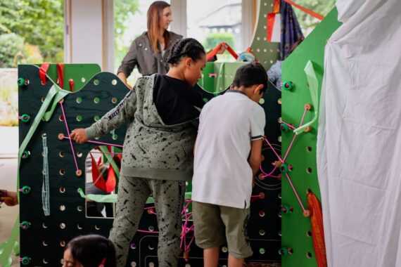 Two children stand at a green pegboard playhouse frame, looping bright pink cords through the holes and securing patterned ribbons; an adult in the further background guides fabric piecesas more cloth panels drape from the interactive structure.