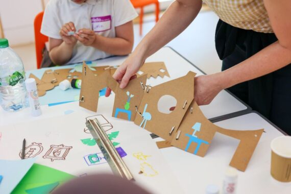A series of cardboard house-shaped panels, stapled together into an elongated strip, each “room” fitted with blue paper cut-outs forming chairs, tables, lamps, and a green vase of flowers. Drawn sketches and glue sticks lie scattered on the table beneath as a child works with a small piece of paper in the background.
