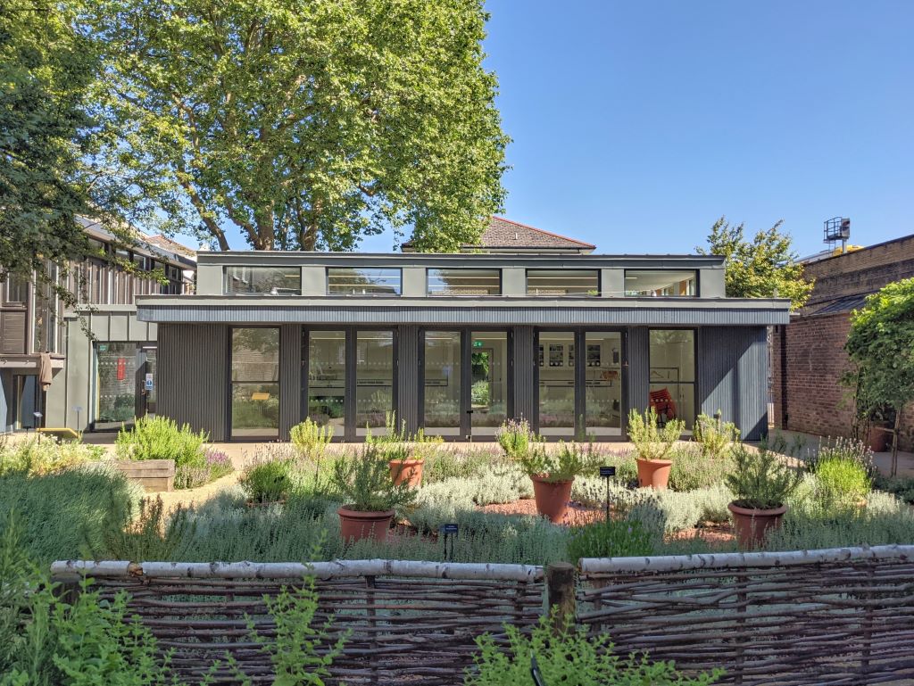 A modern glass-fronted pavilion with dark cladding overlooks a knot garden of potted herbs enclosed by a woven branch fence, bathed in bright sunlight under a clear blue sky.