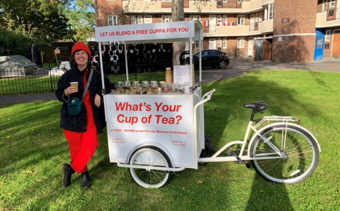 A person in a red beanie and jumpsuit is smiling while holding a paper cup while leaning on a white bicycle cart parked on the grass. The cart’s red lettering matches their outfit, reading “Let us blend a free cuppa for you” above and “What’s Your Cup of Tea?” below, and it’s topped with jars of tea blends.
