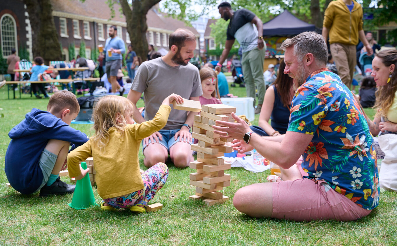 Families playing a large wooden block stacking game on grass at an outdoor community event, with historic brick buildings in the background.
