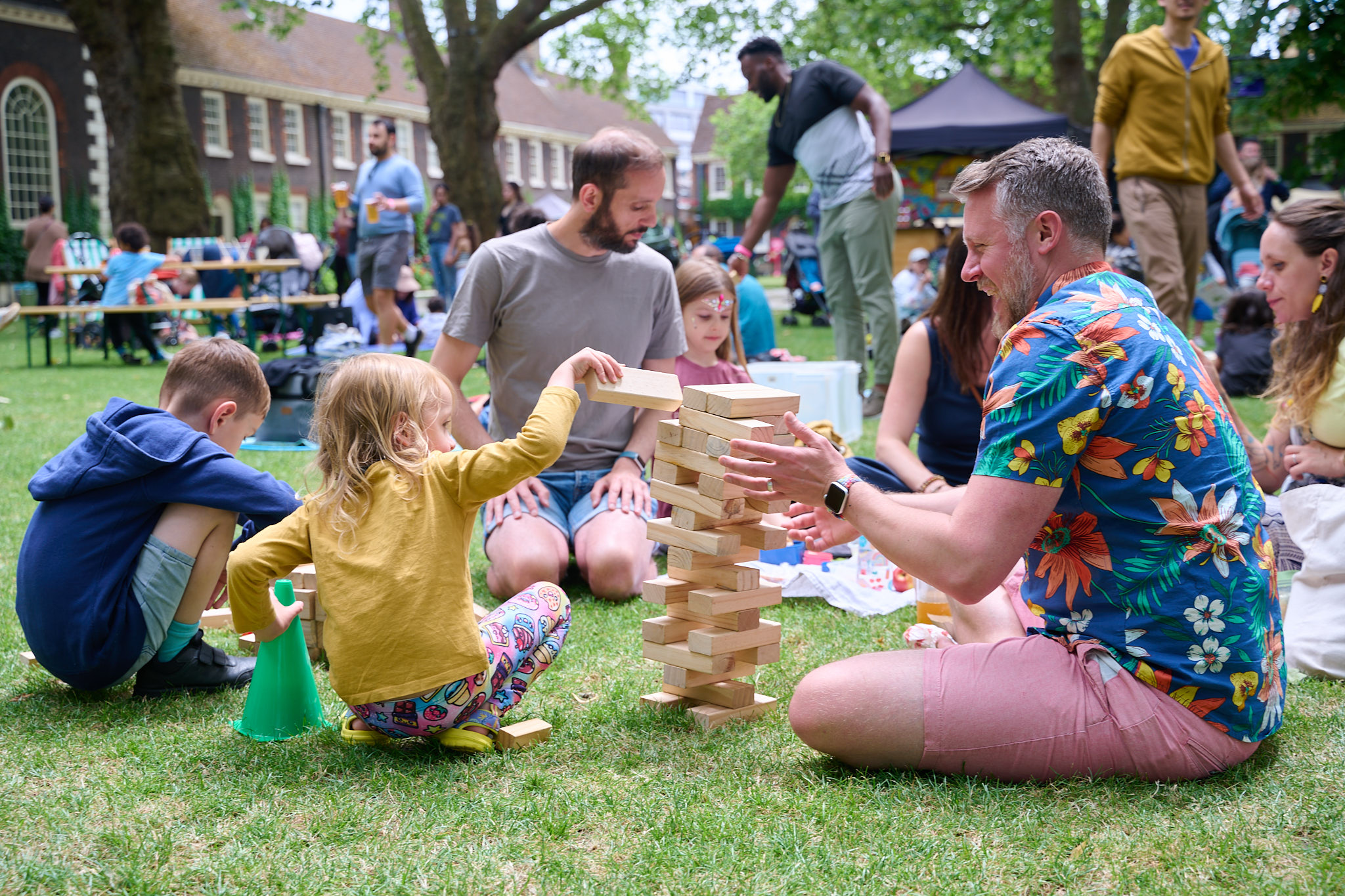 Families playing a large wooden block stacking game on grass at an outdoor community event, with historic brick buildings in the background.