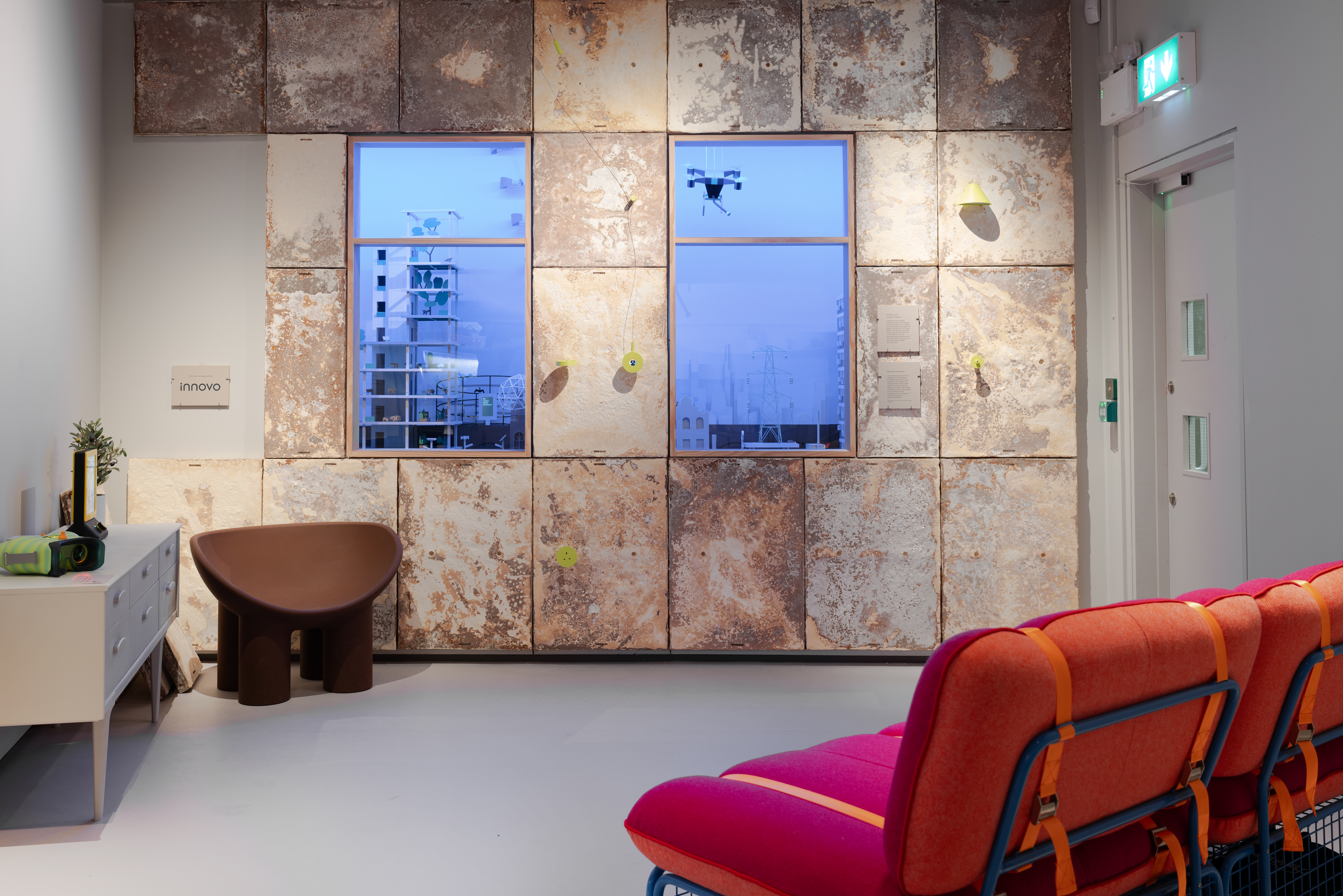 A room with a wall of brown and white panels, two windows showing a futuristic cityscape, and brightly red seating. A brown sculptural chair and a white sideboard with decorative items are also visible.