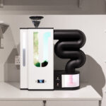 An appliance with a white body and black coiled tubing, set on a counter in a minimalist kitchen. A pink substance is emerging from the tubing.