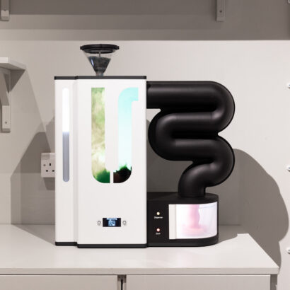 An appliance with a white body and black coiled tubing, set on a counter in a minimalist kitchen. A pink substance is emerging from the tubing.