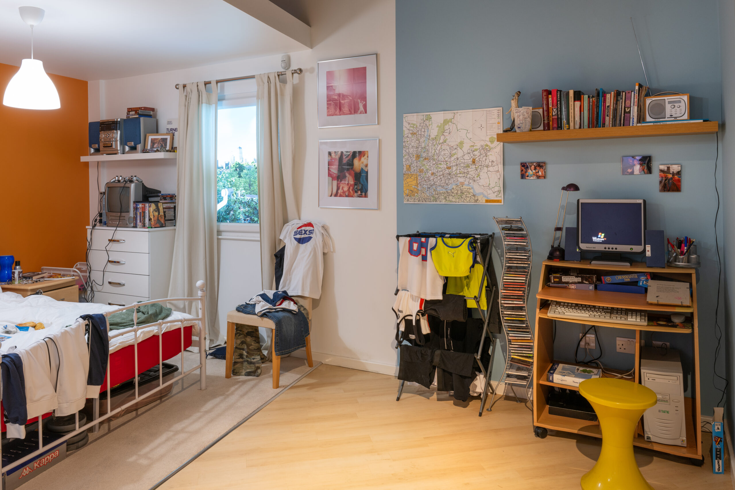 An installation view of the “High-rise Flat in 2005” bedroom: on the left, a vibrant orange wall with white shelves holding a stereo, CRT TV, and DVDs; in front, a white iron-framed bed strewn with clothes. A beige carpet lies beneath. On the right, a pale blue wall displays a map and CDs on narrow shelving above a drying rack of casual wear. Below, a compact wooden desk with keyboard and a CRT monitor showing The Sims, and a bright yellow stool completes the scene.