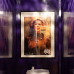 Framed satirical poster in a purple-walled toilet stall above a white cistern: a portrait of Margaret Thatcher with beaded nipple covers and an extravagant bead necklace, captioned “MUZIK – The New Testament of Club Culture” with the Ministry of Sound logo at each bottom corner. Disco-ball reflections shimmer on the purple paint.
