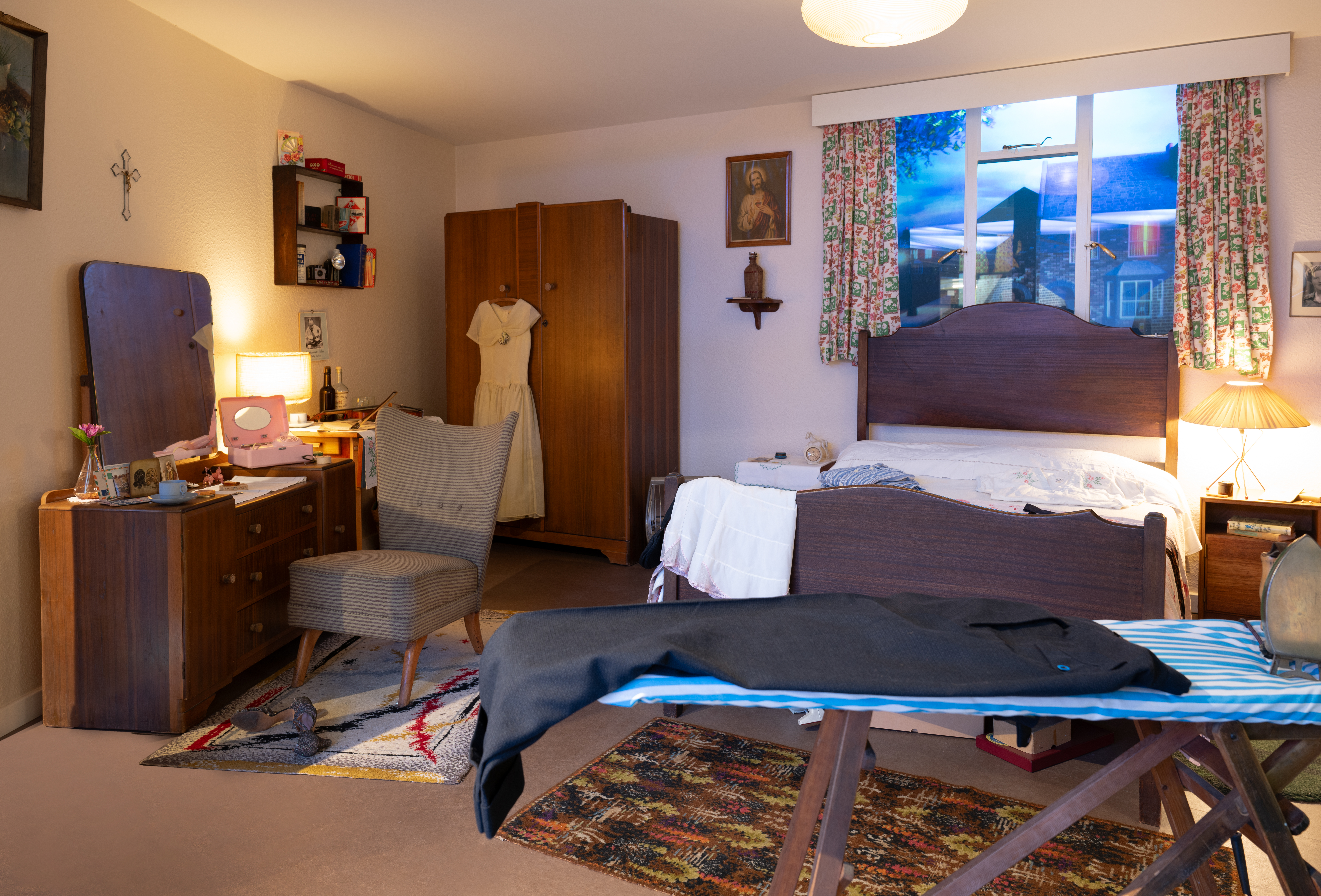 A bedroom featuring dark wood furniture including a bed, wardrobe, and desk. The room has floral curtains, a wooden chair, patterned rugs, and religious artwork on the walls