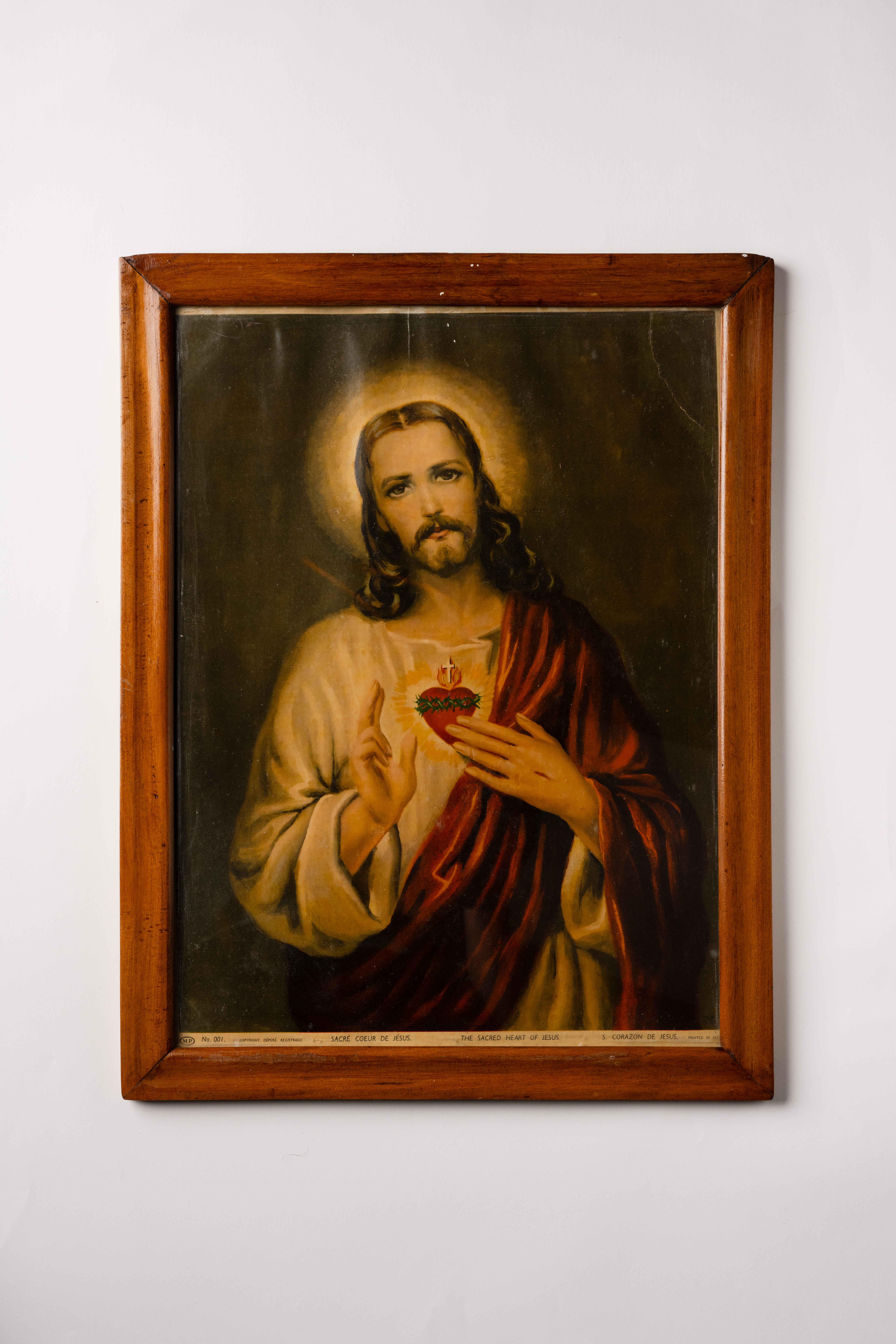 A wooden framed religious artwork showing a figure with long brown hair, a halo, wearing white and red robes, with hands positioned over a glowing heart on the chest..
