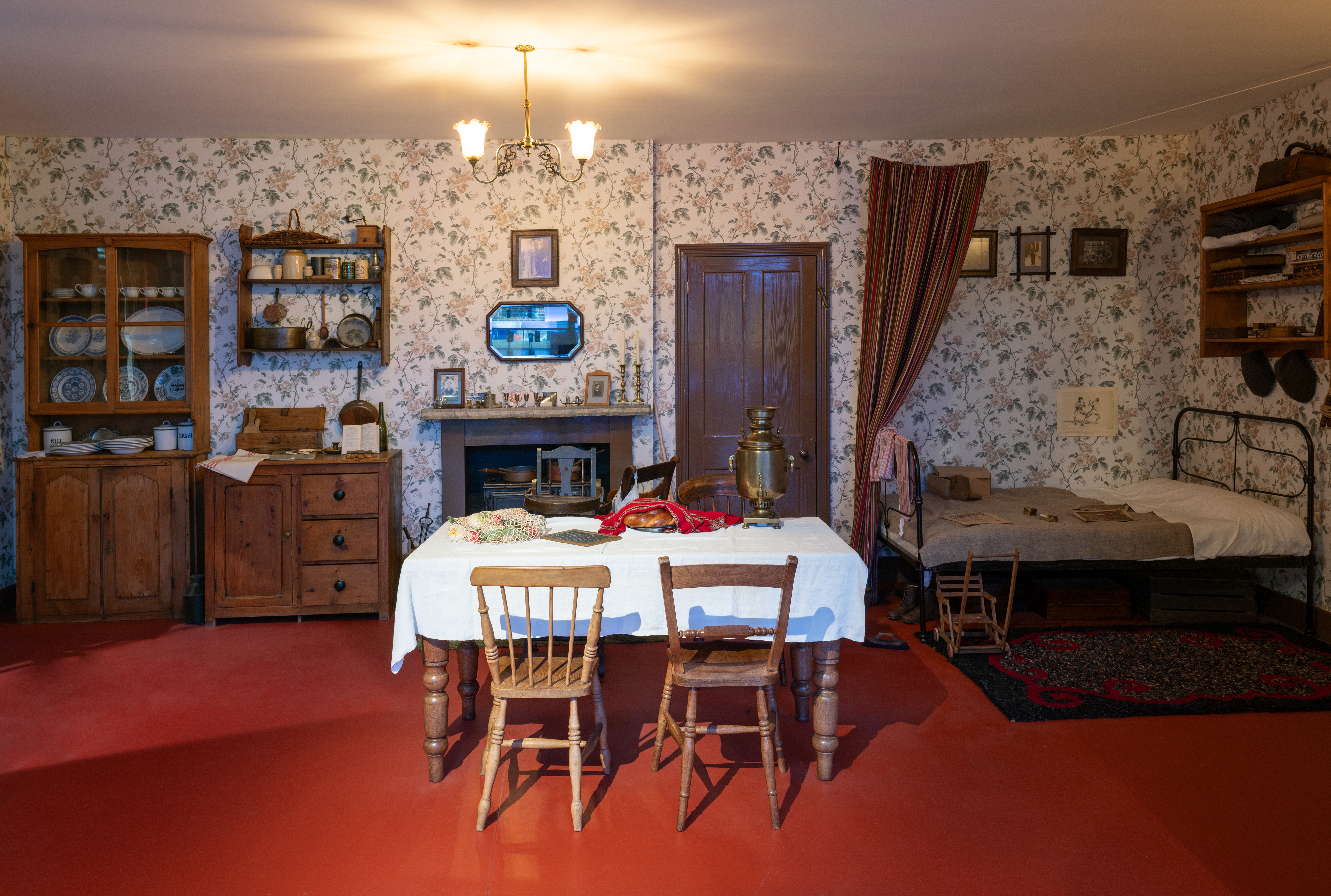 A room with a mantlepiece behind a wooden dining table laid with a white tablecloth and two wooden chairs. To the right a colourful curtain reveals a brass bed frame is visible, and to the left wooden storage with various dishware and china.