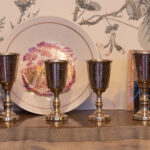 Four silver Kiddush cups arranged in front of a decorative plate with floral wallpaper background.