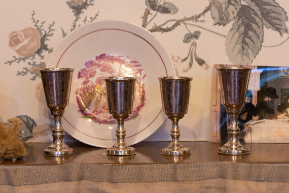 Four silver Kiddush cups arranged in front of a decorative plate with floral wallpaper background.