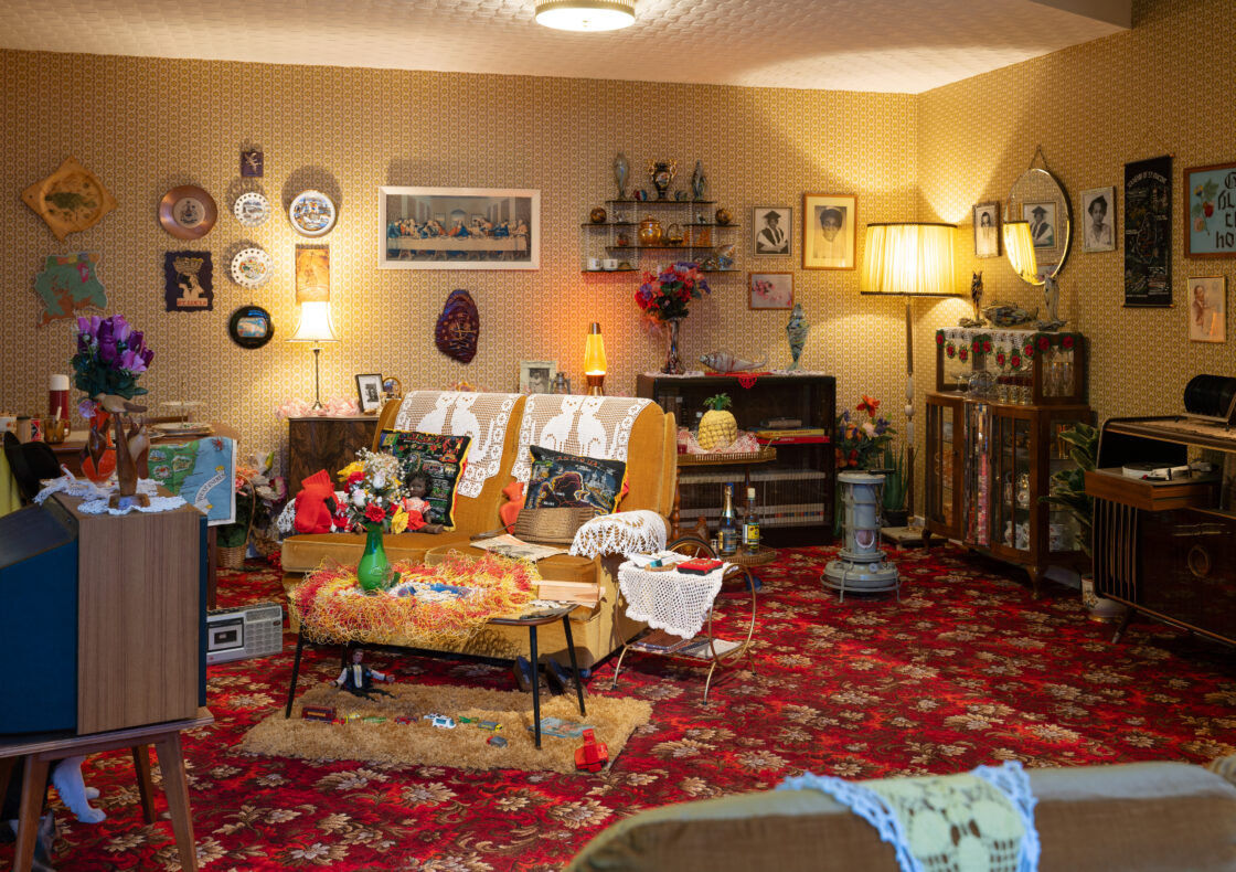 A cluttered 1970s-style living space with yellow wallpaper, red patterned carpet, and furniture and decorations.
